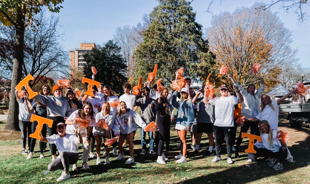 Students cheering on a sunny campus lawn with orange T signs and pom-poms