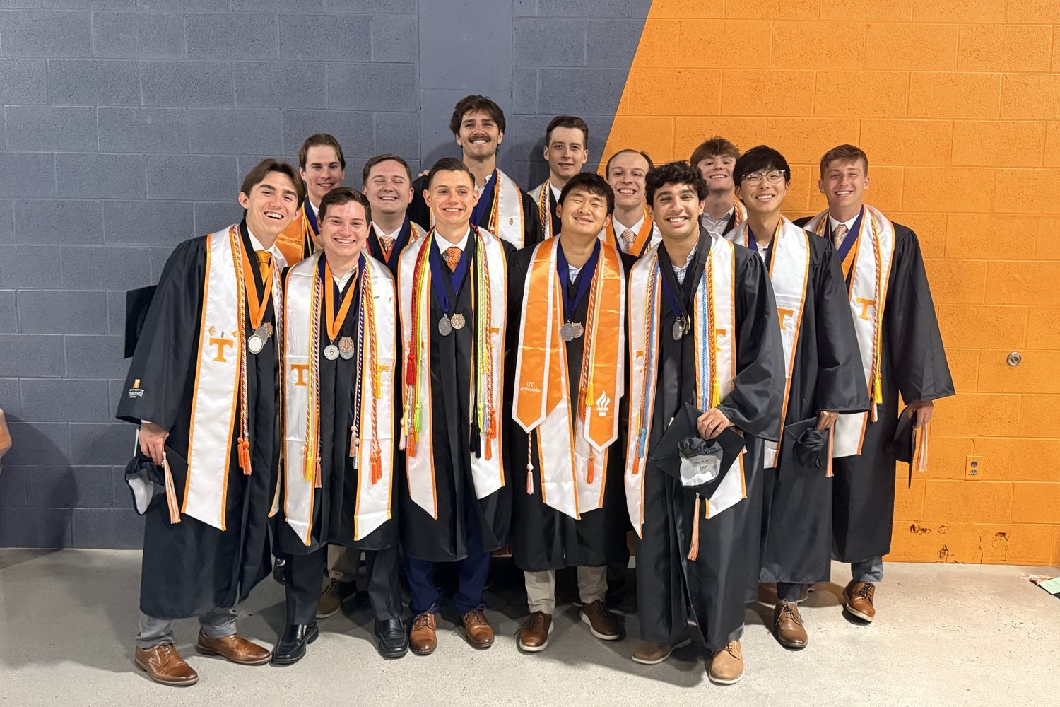 Smith GLS graduates in caps, gowns and honor stoles in front of an orange and gray wall
