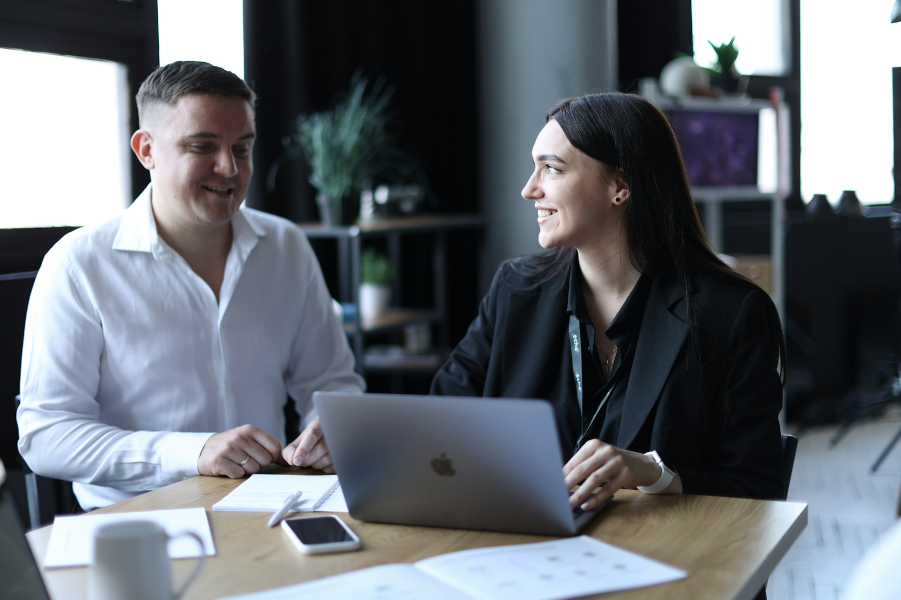 Two colleagues at a table turned toward each other, smiling and listening closely