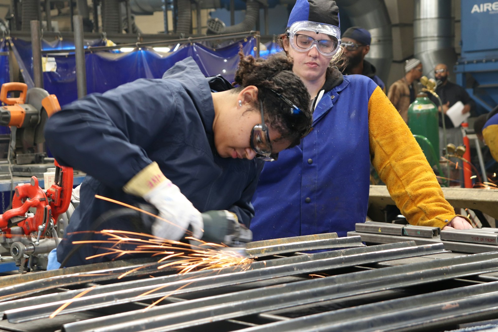 An instructor guiding an apprentice through metalwork in a training shop