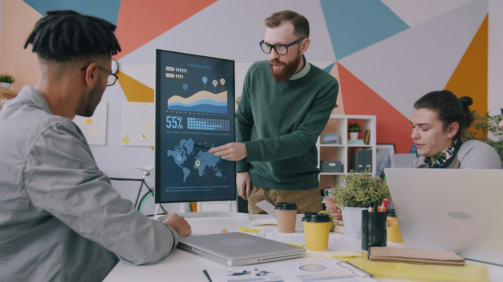 A man pointing at a vertical monitor of data charts while two colleagues look on