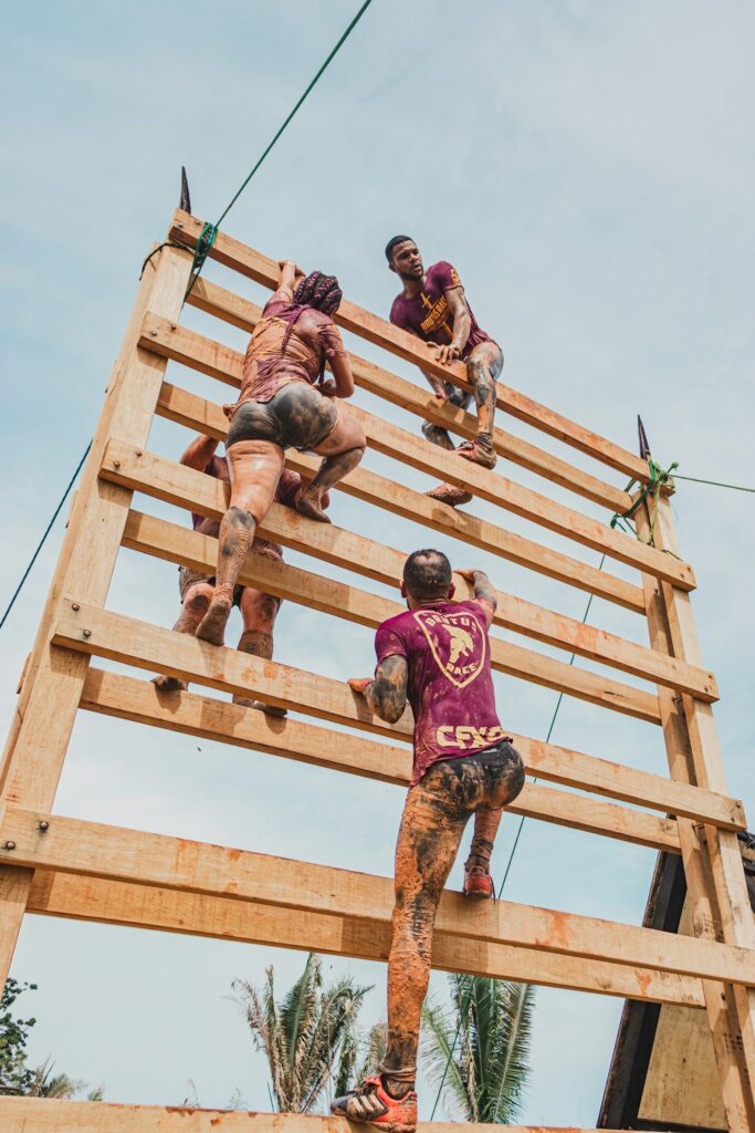 Three people helping each other climb a tall wooden obstacle wall