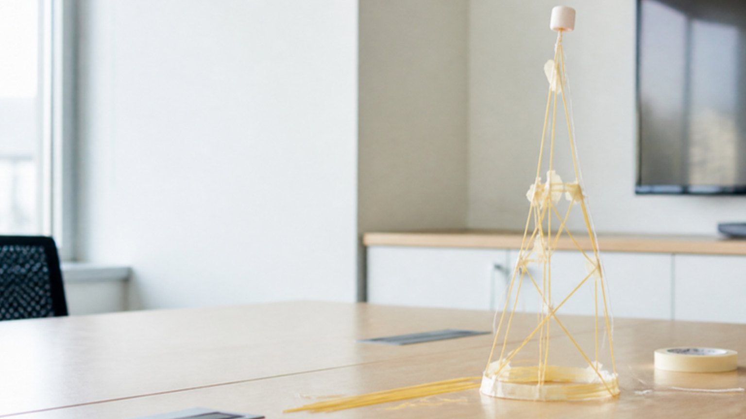 A tower of spaghetti, tape and string on a conference table with a marshmallow balanced on top