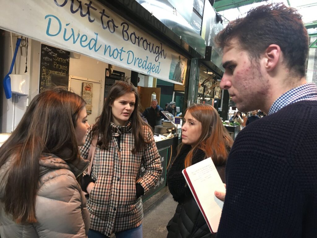 Students take notes while talking with a stranger outside a London market stall